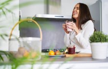 Jovem rindo mulher segurando caneca e tomando café da manhã na cozinha moderna em casa — Fotografia de Stock