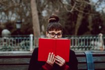 Joven mujer alegre en anteojos mirando a la cámara y cubriendo la cara con libro mientras está sentado en el banco en el jardín de la ciudad - foto de stock