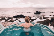 Vista trasera de la mujer en traje de baño descansando en el agua de la piscina cerca de rocas y cielo nublado en la costa del mar - foto de stock