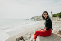 Stylische Frau schaut weg, während sie auf einem Felsen am Strand sitzt — Stockfoto