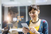 Manager femminile sorridente utilizzando smartphone mentre in piedi vicino alla parete di vetro in ufficio moderno — Foto stock