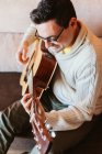 Sorrindo homem em óculos tocando guitarra no sofá em casa — Fotografia de Stock