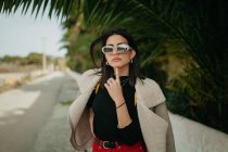 Stylish young woman in sunglasses standing near tropical palm leaves on street — Stock Photo