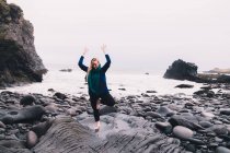 Jovem mulher com as mãos levantadas meditando e de pé em rochas perto da costa do mar — Fotografia de Stock
