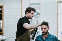 Barber applying hair gel on hair of handsome stylish male sitting in chair in barbershop — Stock Photo