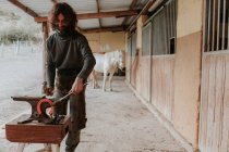 Guerrier adulte utilisant un marteau et des pinces pour forger un fer à cheval chaud sur une enclume portable près d'une écurie sur un ranch — Photo de stock