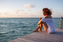 Seitenansicht einer hübschen jungen Frau, die auf einem hölzernen Pier in der Nähe von gewelltem Meerwasser vor wolkenverhangenem Abendhimmel auf den Malediven sitzt — Stockfoto