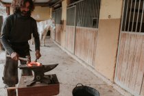 Erwachsener Hufschmied schmiedet mit Hammer und Zange heißes Hufeisen auf tragbarem Amboss in der Nähe des Stalls auf der Ranch — Stockfoto