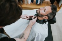 Peluquero con peine y recortadora barba de corte de macho sentado en la barbería sobre fondo borroso - foto de stock