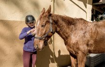 Nettes kleines Mädchen mit Helm beim Putzen eines weißen Pferdes, während es in der Nähe von Ställen im Stall während des Reitunterrichts auf der Ranch steht — Stockfoto