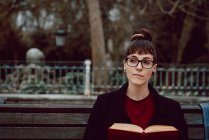 Молода елегантна жінка в окулярах з книгою, що сидить на лавці в міському парку — стокове фото