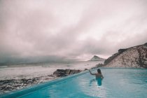Back view of young woman upped hand resting in water of pool near cliffs on coast and stormy sea — Stock Photo