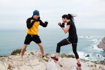 Uomo e donna in guantoni da boxe che si picchiano a vicenda mentre stanno in piedi sulla scogliera contro mare e cielo — Foto stock