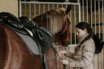 Vista lateral da jovem fêmea no capacete colocando sela no cavalo maravilhoso perto estável no rancho — Fotografia de Stock