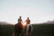 Visão traseira do homem e da mulher montando cavalos e segurando as mãos contra o céu pôr do sol na fazenda — Fotografia de Stock