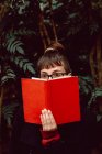 Young elegant woman in eyeglasses looking away while covering face with book in city garden — Stock Photo
