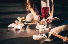 Crop women sitting on wooden floor putting on satin pointe shoes in sunlight. — Stock Photo