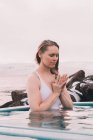 Молодая женщина с закрытыми глазами медитирует в воде бассейна возле скал и облачного неба — стоковое фото