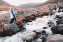Jovem mulher com as mãos erguidas meditando e em pé sobre rochas perto de estreito rio rápido entre colinas — Fotografia de Stock