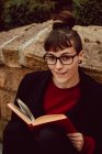 Junge elegante lächelnde Frau mit Brille lehnt mit Buch an Steinmauer und blickt in die Kamera — Stockfoto