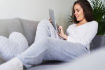 Junge glückliche Frau nutzt digitales Tablet und ruht sich zu Hause auf dem Sofa aus — Stockfoto