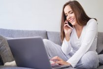 Усміхнена молода щаслива жінка використовує ноутбук і розмовляє на мобільному телефоні на дивані вдома — стокове фото