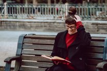Молода усміхнена елегантна жінка в окулярах читає книгу і сидить на лавці в міському парку — стокове фото