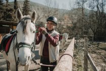 Macho sênior no capacete colocando freio no cavalo branco durante a aula de equitação no dia de outono no rancho — Fotografia de Stock