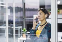 Жіночий менеджер розмовляє на мобільному телефоні, стоячи біля скляної стіни в сучасному офісі — стокове фото