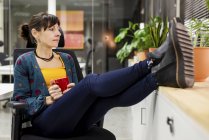 Задумчивая женщина-менеджер с кружкой горячего напитка отдыхает на стуле в офисе — стоковое фото