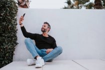 Edad media guapo elegante hombre feliz en ropa casual tomar selfie en el teléfono móvil y sentado cerca de la pared gris - foto de stock