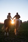 Vue arrière de l'homme et de la femme chevauchant des chevaux et se tenant la main contre le ciel couchant sur ranch — Photo de stock