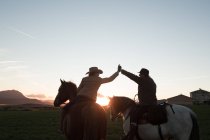 Vista trasera del hombre y la mujer montar a caballo y dar cinco de altura entre sí contra el cielo puesta del sol en el rancho - foto de stock