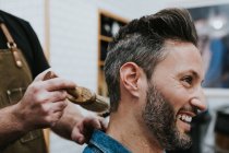 Side view of crop barber with brush near happy handsome stylish male sitting in chair in salon — Stock Photo
