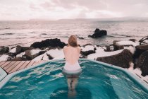 Vista traseira da mulher em maiô descansando na água da piscina perto de rochas e céu nublado na costa do mar — Fotografia de Stock