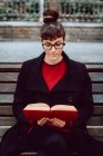 Jeune femme élégante attrayante dans le livre de lecture de lunettes et assise sur le banc dans le jardin de la ville — Photo de stock