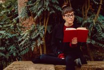 Joven mujer elegante en gafas de lectura libro y sentado en el banco en el jardín de la ciudad - foto de stock
