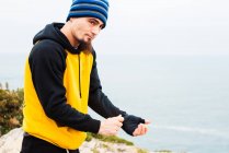 Бородатий дорослий чоловік упаковка пов'язку навколо руки, стоячи проти моря під час активного навчання з кікбоксингу — стокове фото