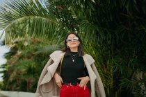 Elegante giovane donna in occhiali da sole in piedi vicino a foglie di palma tropicale sulla strada — Foto stock