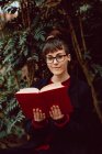 Giovane donna elegante attraente in occhiali da vista con libro e seduto sulla panchina in giardino della città e guardando la fotocamera — Foto stock