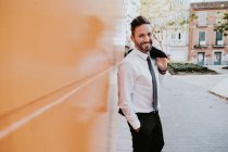 Vista lateral do adulto bonito elegante empresário alegre em terno formal com mão no bolso olhando para a câmera perto da parede laranja na rua da cidade — Fotografia de Stock