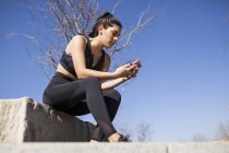 Брюнетка женщина сидит на бетонной стене и проверяет статистику тренировочного приложения — стоковое фото