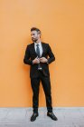 Adulto guapo elegante hombre de negocios en traje formal ajustando la chaqueta y mirando hacia otro lado cerca de la pared naranja - foto de stock