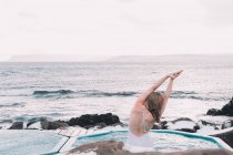 Вид сзади женщины в купальнике, отдыхающей в воде бассейна рядом со скалами и облачным небом на морском побережье — стоковое фото