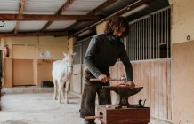 Взрослый кузнец, использующий молоток и щипцы для ковки горячей подковы на переносном наковальне возле конюшни на ранчо — стоковое фото