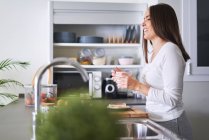 Вид сбоку на молодую привлекательную женщину, держащую кружку на современной кухне дома — стоковое фото