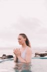 Junge Frau mit geschlossenen Augen meditiert im Wasser des Pools in der Nähe von Felsen und bewölktem Himmel — Stockfoto