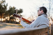 Casual man in eyeglasses playing guitar on bench in countryside — Stock Photo
