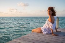 Seitenansicht einer hübschen jungen Frau, die lächelt und in die Kamera blickt, während sie auf einem hölzernen Pier in der Nähe von plätscherndem Meerwasser vor bewölktem Abendhimmel auf den Malediven sitzt — Stockfoto