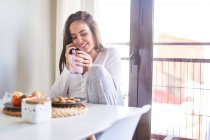 Joven feliz mujer hablando en el teléfono móvil mientras desayuna en la mesa cerca de la ventana en casa - foto de stock